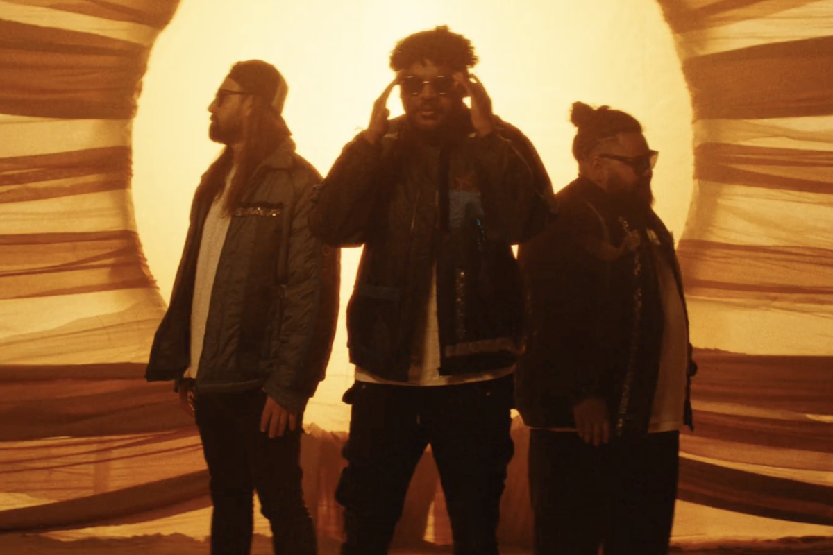 First Nations hip-hop group DEM MOB standing together silhouetted against a bright glowing circle backlighting draped fabric in warm golden hues.